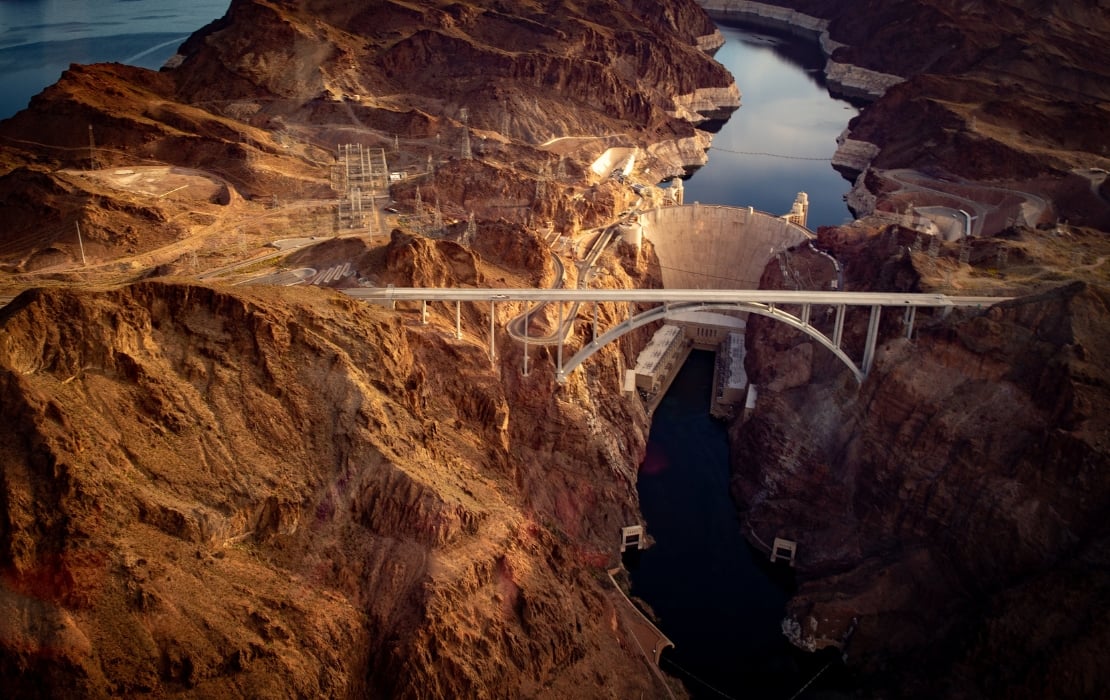 An aerial photo of the Hoover Dam in the Black Canyon of the Colorado River. The dam is just behind the Colorado River Bridge. The canyon is very prominent in the image, with the red rock standing out.