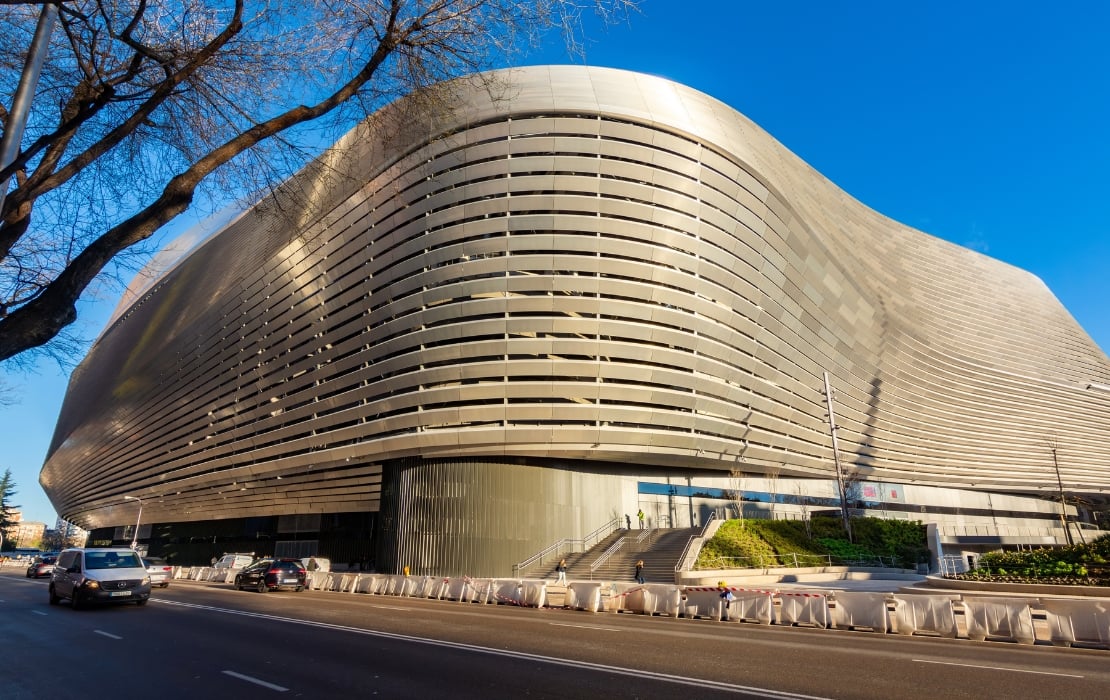 A photo of the exterior of the renovated Santiago Bernabéu Stadium in Madrid, Spain, featuring its new steel facade, made of 7,500 V-shaped slats. It stands out against a bright blue sky.