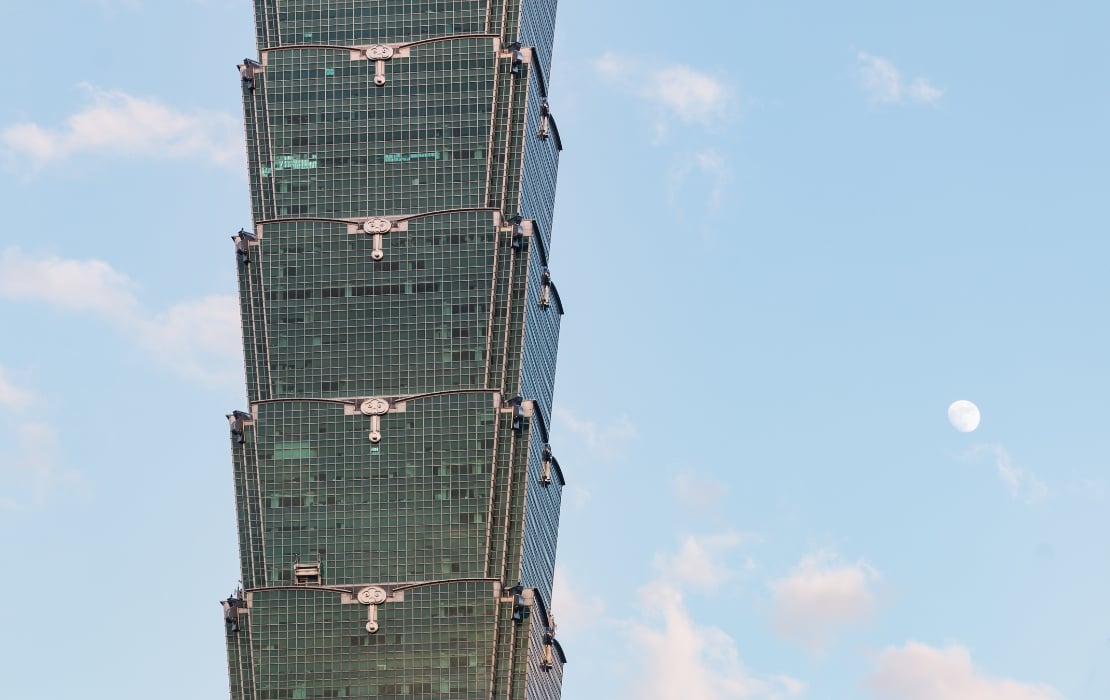 Close-up view of the tiered glass façade of Taipei 101, with a pale blue sky and the moon visible to the right.