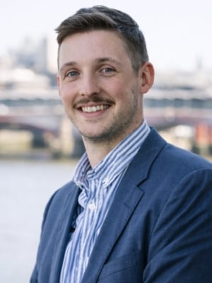 Head-and-shoulders portrait of a person wearing a blue suit jacket and striped shirt, photographed outdoors with a river and bridge visible in the blurred background.