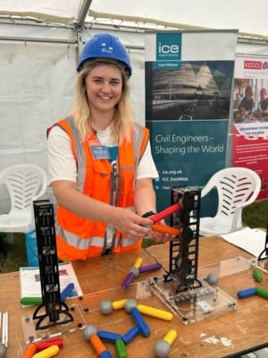 Person wearing a hard hat and high-visibility vest demonstrating a model bridge-building activity at an ICE STEM outreach event.