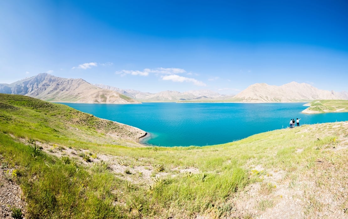 Panoramic view of Lar Dam in Iran with turquoise water surrounded by green hills and rugged mountains under a clear blue sky.
