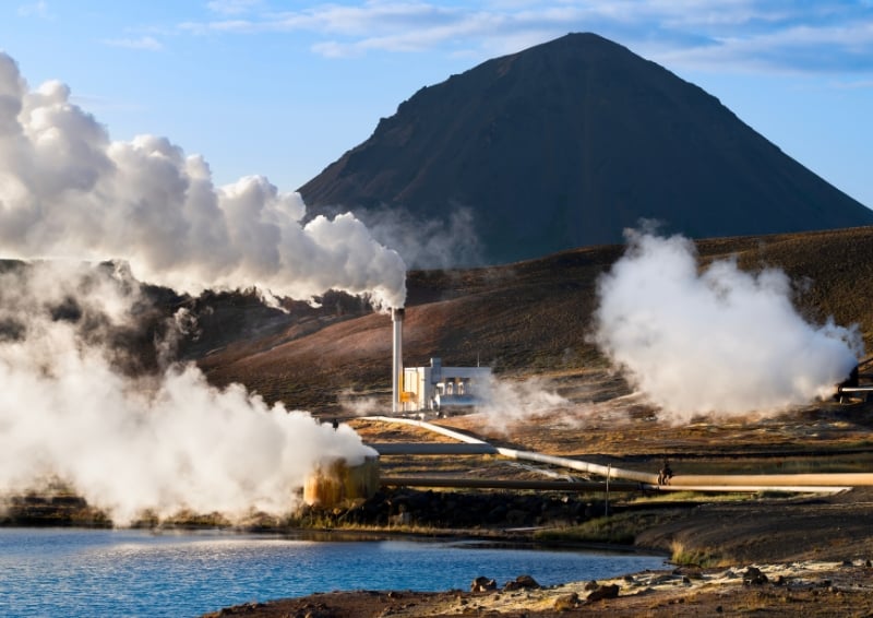 Geothermal power plant with steam rising from multiple vents and pipes beside a lake, set against a rugged volcanic landscape with a large mountain in the background under a partly cloudy sky.