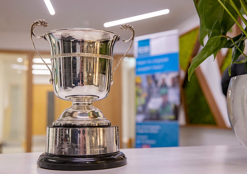 Polished silver trophy cup with decorative handles mounted on a dark base, displayed on a table in a modern office setting, with blurred signage and a potted plant in the background.