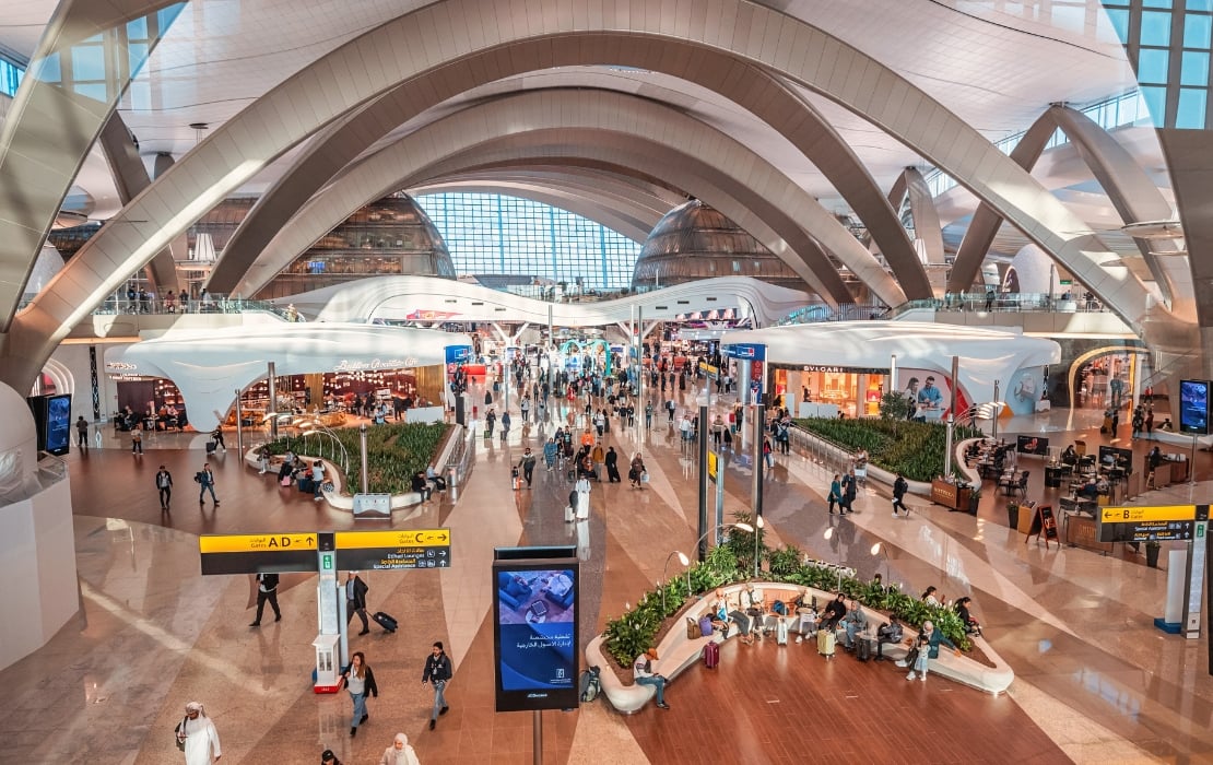 Interior view of Abu Dhabi International Airport terminal with modern curved architecture, busy shopping area, and travelers walking through spacious concourse.