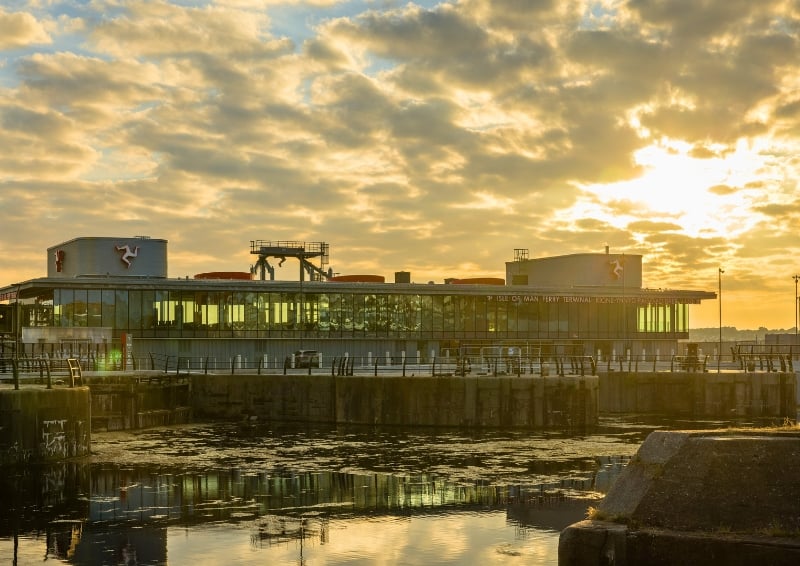 Modern glass-fronted ferry terminal building on a waterfront at sunset, with golden sunlight reflecting off calm water in the foreground and dramatic clouds overhead.
