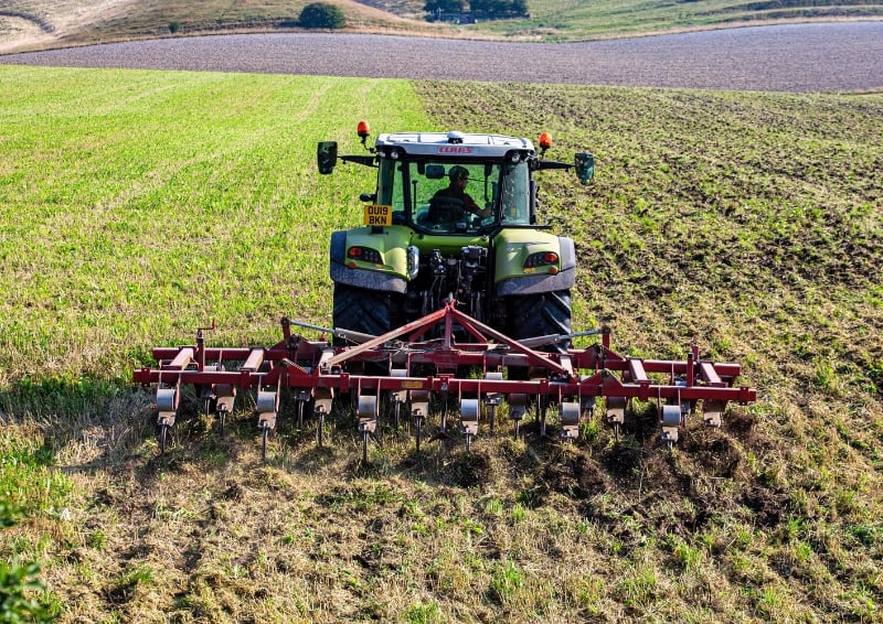 Tractor with a rear-mounted cultivator working across a field, turning soil between strips of green crop and bare earth in a rural landscape.