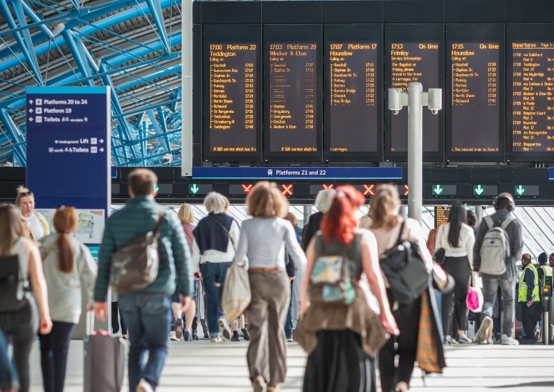 People walking through a busy railway station concourse beneath large digital departure boards showing train times and platforms.