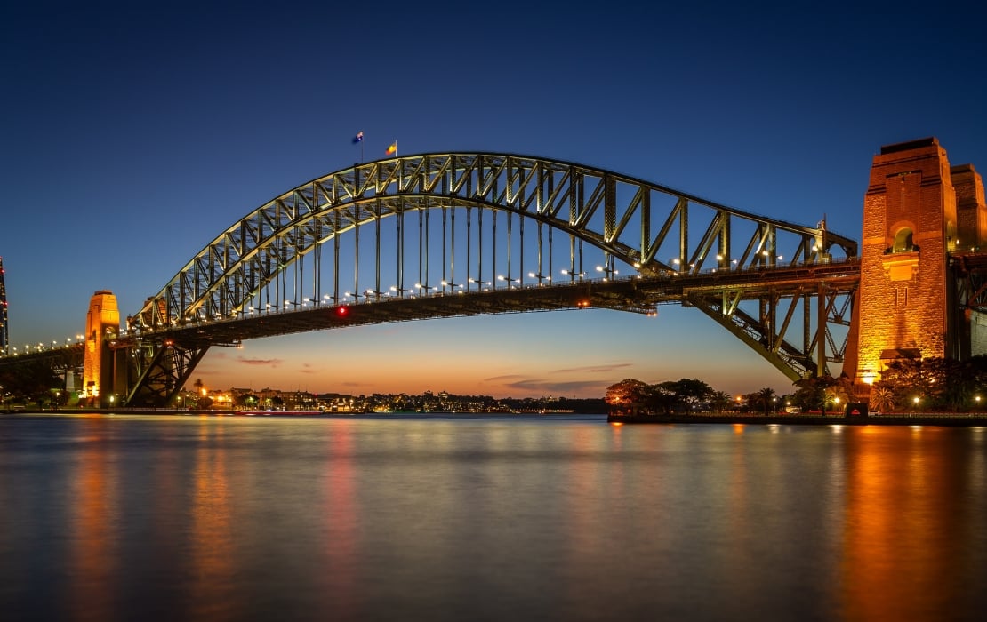 A photo of the Sydney Harbour Bridge at twilight, with the sun setting in the background and a night sky descending. The bridge's steel arch design is lit up by a few lights throughout its span and on either side.