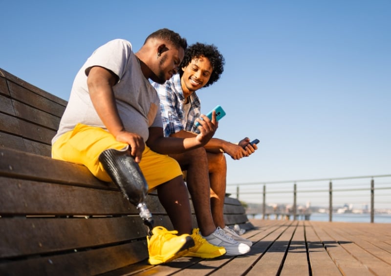 Two men sitting on a wooden bench outdoors near a waterfront, both holding smartphones. One man is wearing a gray T-shirt, bright yellow shorts, and yellow sneakers, with a visible prosthetic leg. The other man is wearing a checkered shirt and shorts.