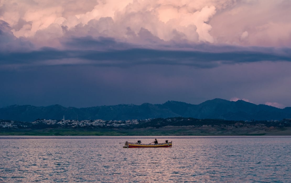 Fishing boat floating on Mangla Dam reservoir in Pakistan with distant hills and dramatic cloudy sky at sunset.