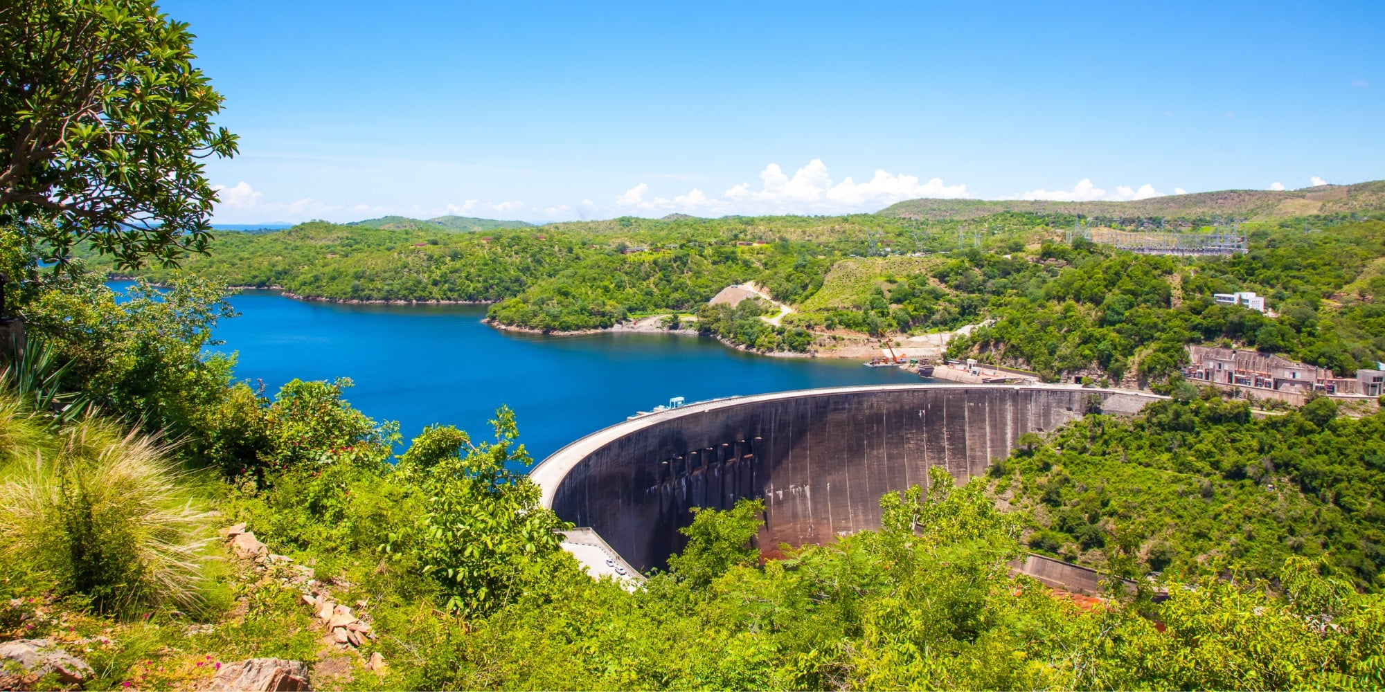 A photo of an aerial view of the Kariba dam, an arched structure. The water in the reservoir is vibrant blue and it's surrounded by trees.