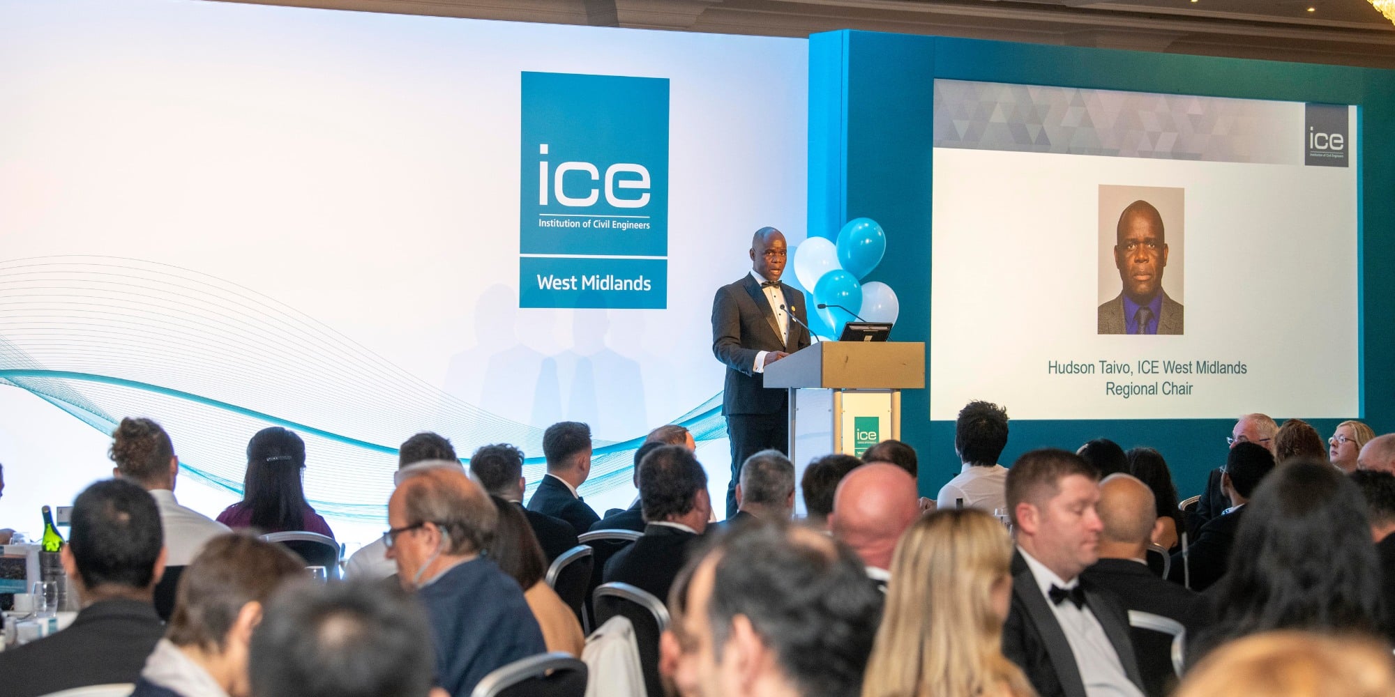 A photo of Hudson Taivo speaking at a podium at the ICE West Midlands Awards ceremony. He is on a stage and there is a presentation slide behind him with his picture and name. In the foreground, there is a seated audience around round tables. 