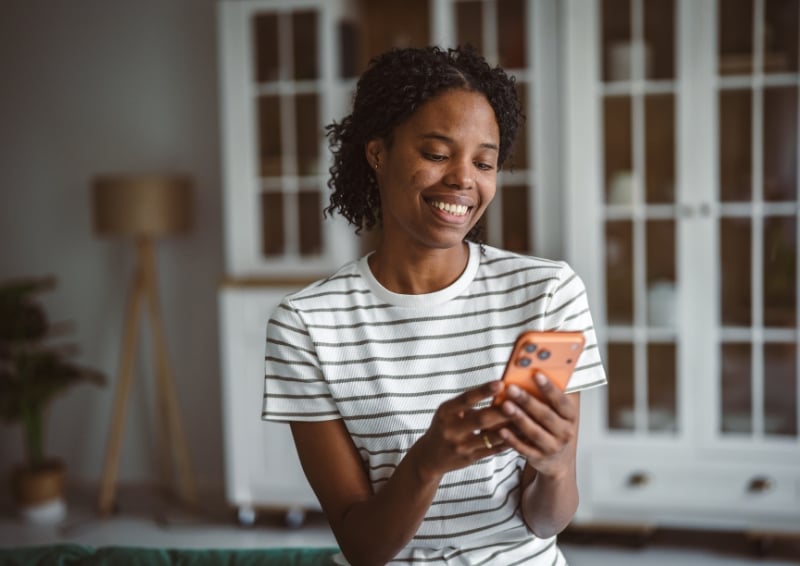 Black woman sitting indoors and using a smartphone, wearing a striped T-shirt, with a softly lit room and cabinet furniture in the background