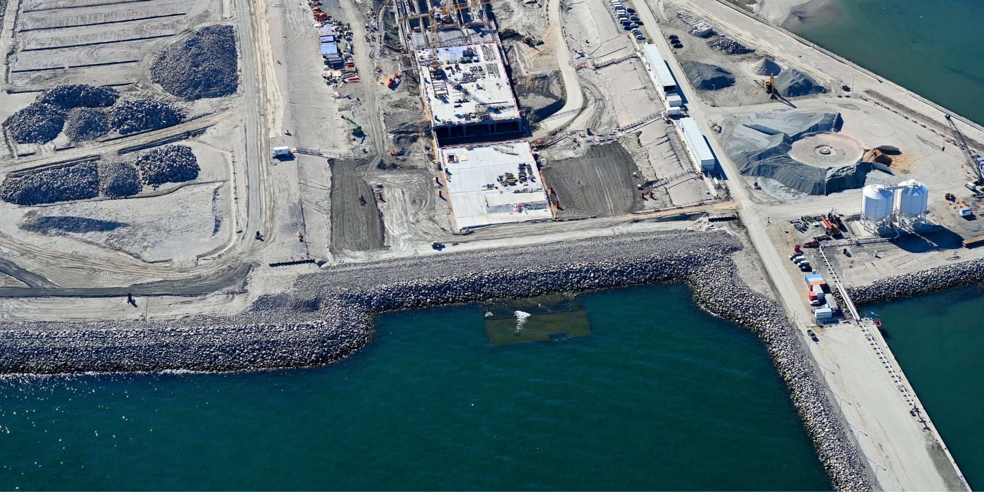 An aerial image of the German construction site on the island of Fehmarn. The top half of the image shows the construction site with some vehicles onsite. The bottom half is the deep-blue green of the sea. Image copyright: Sund & Bælt Holding A/S