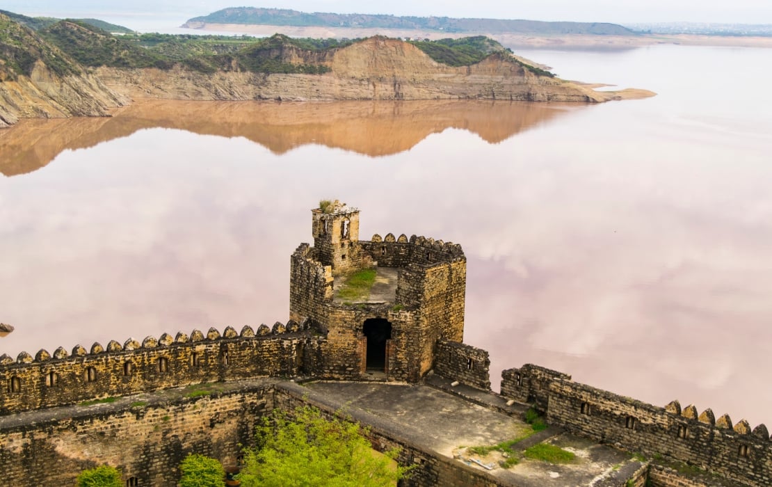 Historic fort overlooking Mangla Dam reservoir with calm water and rugged hills in the background.