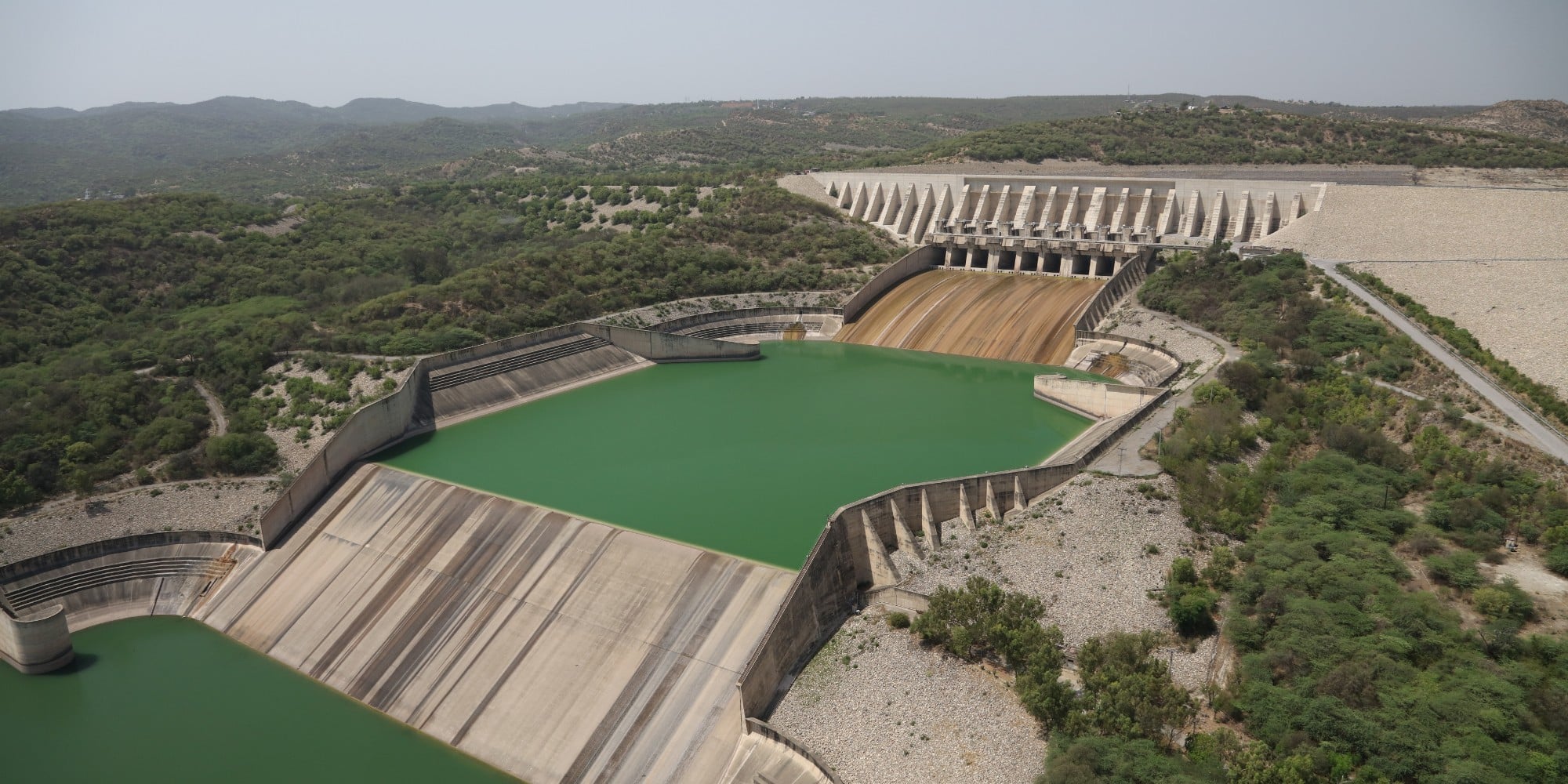 Aerial view of Mangla Dam with green reservoir water, spillway gates, and surrounding hills in a hydroelectric and irrigation project setting.