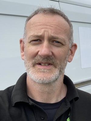Head-and-shoulders portrait of a person with short grey hair and a beard, wearing a black polo shirt and photographed outdoors against a light-coloured background.
