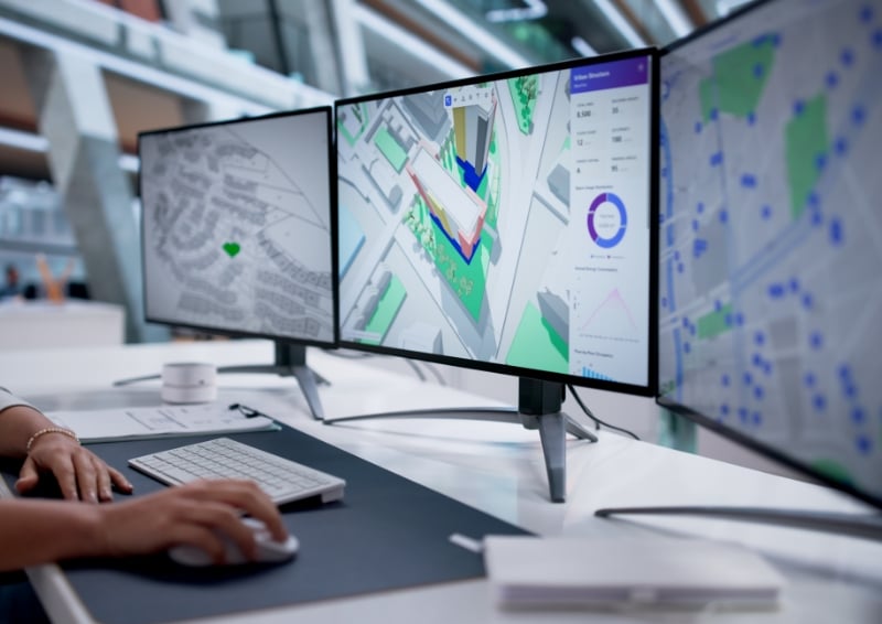 Three computer monitors on a desk display detailed 3D building models and map-based data dashboard
