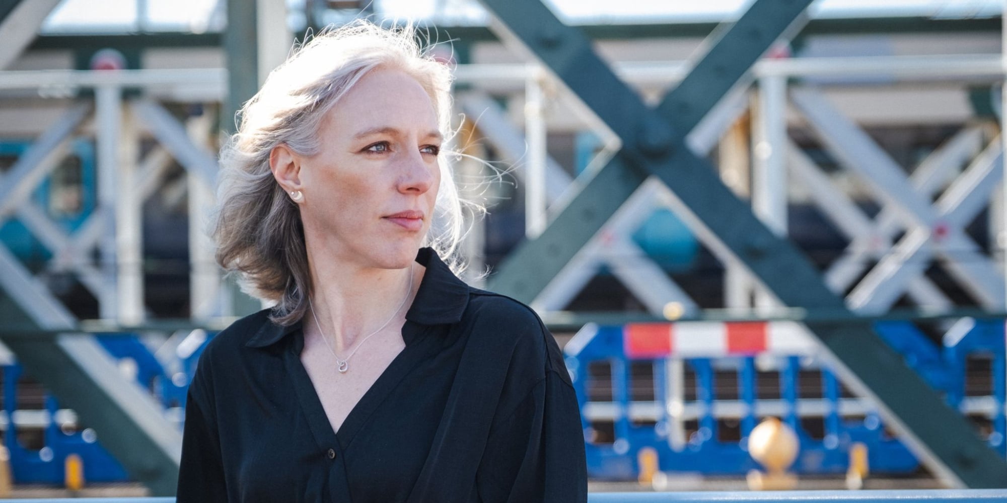Andrea Charlson wearing a black collared shirt stands outdoors in front of a metal structure with crisscross beams. The background includes industrial elements and blue machinery, with sunlight illuminating the scene.