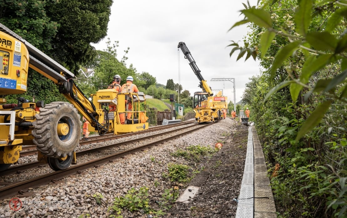 A photo of plant machinery working on a South Wales Metro site, driving over the tracks of the railway. On either side of the yellow machines, there are trees.