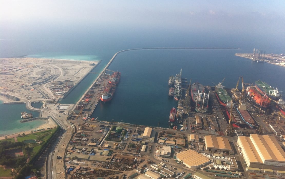 Aerial view of Port Rashid in Dubai, showing cargo ships docked at the harbor, industrial facilities, and surrounding coastline with turquoise waters.