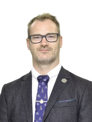 A portrait of Alex Soles wearing a dark grey suit