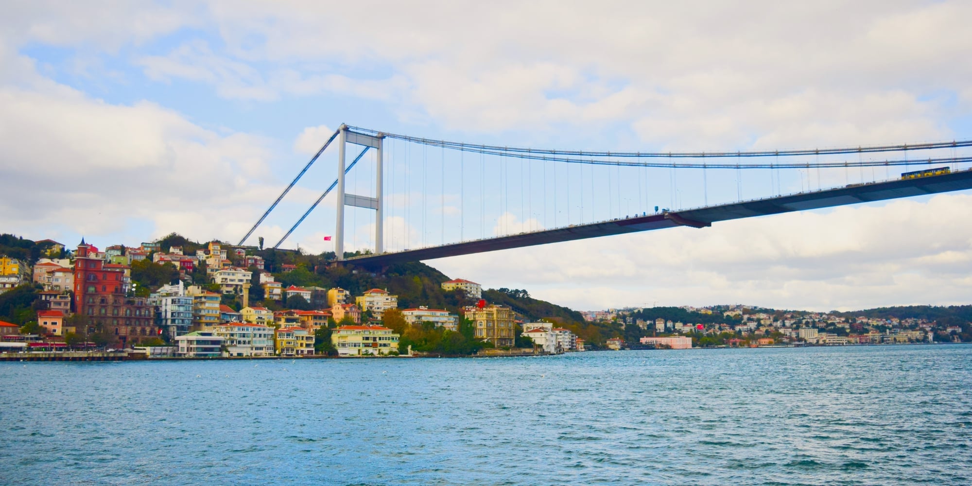 Suspension bridge stretching across a wide waterway under a partly cloudy sky, with colorful hillside buildings and greenery along the shoreline.