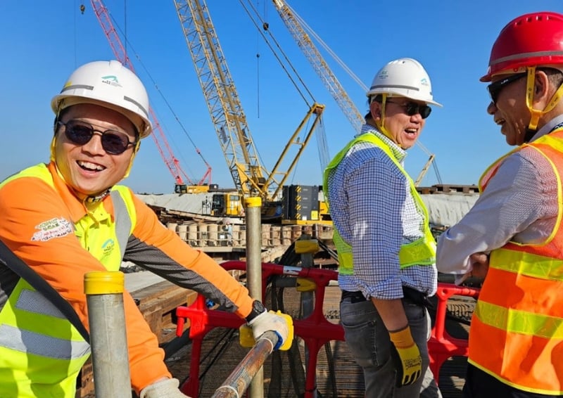 Three construction site workers wearing hard hats, high-visibility vests, and gloves stand and lean on a railing in the foreground, with large cranes and active building works visible in the background under a clear blue sky.