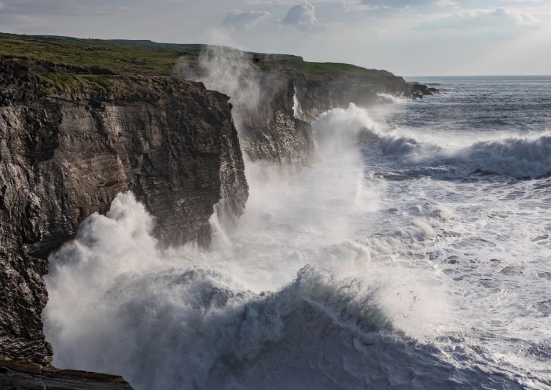 Large waves crash powerfully against tall, rugged sea cliffs under a bright, partly cloudy sky. 