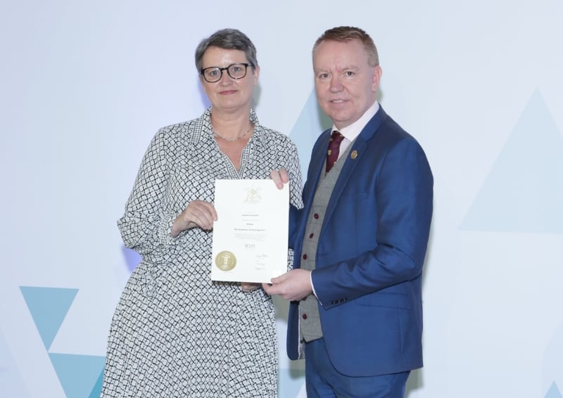 Sue Caccavone and David Porter standing side by side, holding an official certificate, posed against a light backdrop with geometric shapes.