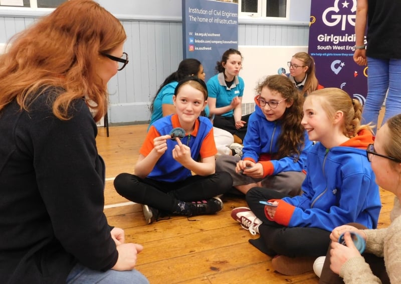 A group of young people in brightly coloured uniforms sit on a wooden floor in a circle, participating in an activity and holding small objects