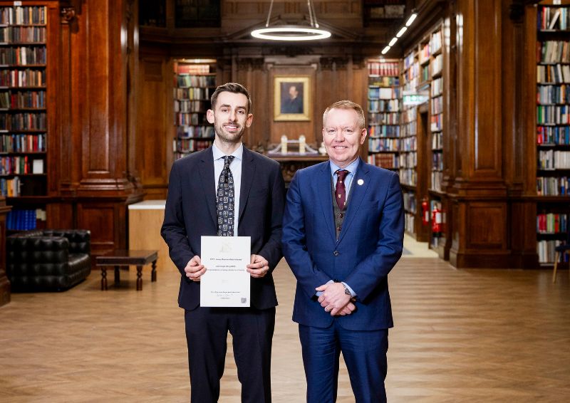 Liam Eagle and David Porter stand side by side in an ornate library with tall wooden bookshelves filled with books. 